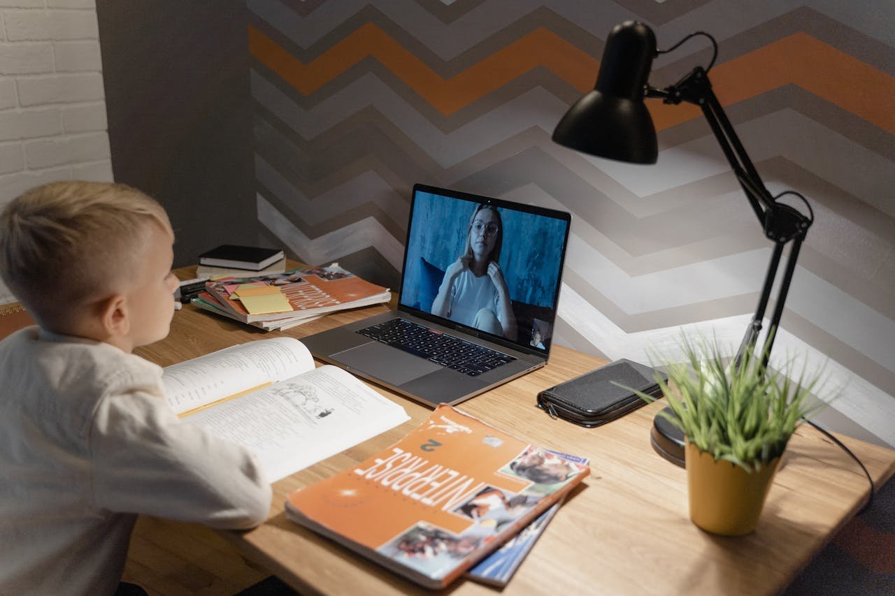 A young boy participates in a virtual class from home, using a laptop and study materials.